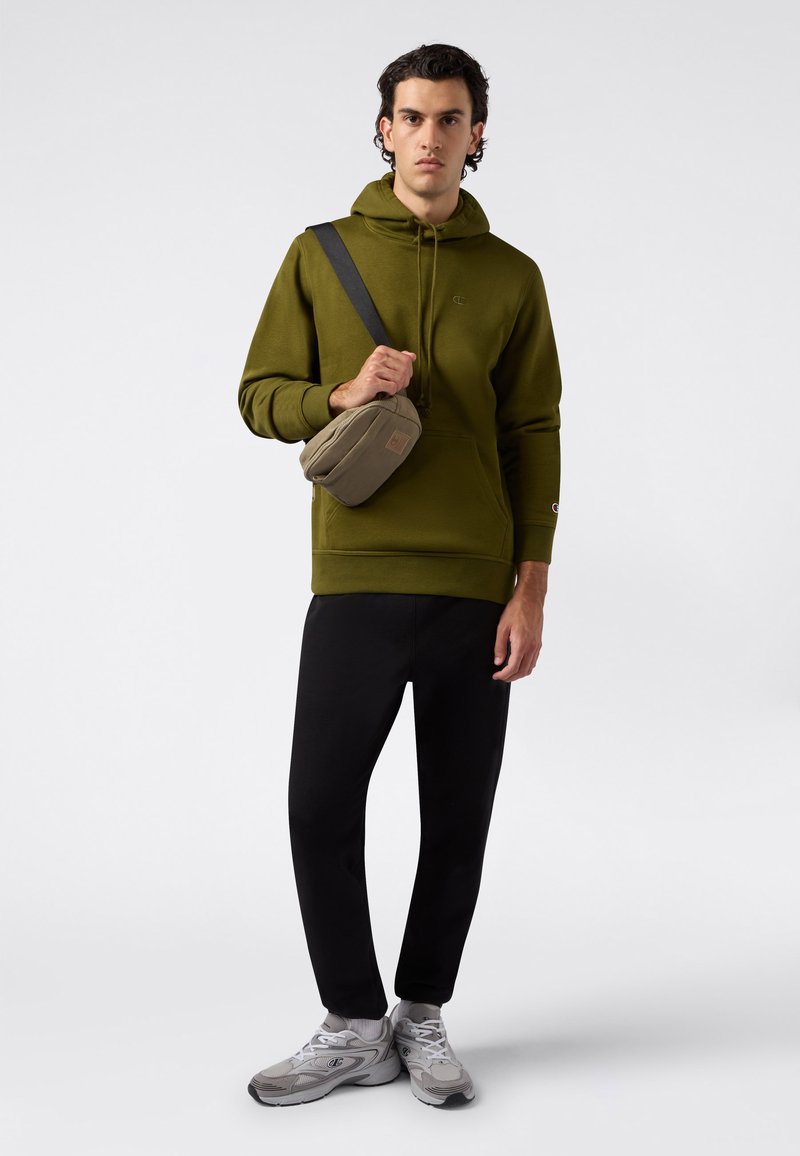 Mężczyzna w oliwkowym kapturze, czarnych spodniach, szarych sneakersach i beżowej torbie na ramię na tle białym.