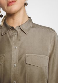 Camicia bottone color khaki con colletto, dotata di due tasche frontali, texture liscia e chiusure a bottone trasparenti.