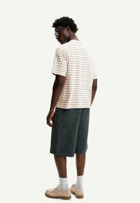 Striped short-sleeve t-shirt in light colors, paired with dark gray shorts, and beige shoes. The model poses from the back, wearing sunglasses.