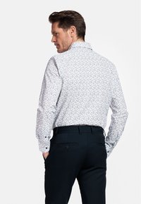 Chemise blanche à manches longues avec un motif floral multicolore ; associée à un pantalon sombre ; boutons visibles aux poignets ; coupe ajustée.