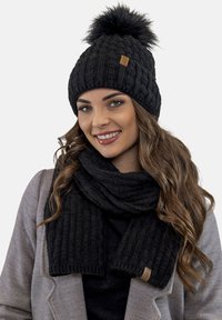Berretto lavorato a maglia nero con pom-pom e sciarpa nera coordinata. Entrambi presentano un motivo testurizzato e un piccolo etichetta con logo marrone.