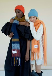 Deux modèles portent des écharpes surdimensionnées et des bonnets tricotés. La première écharpe présente un motif multicolore, tandis que la seconde est unie et orange.
