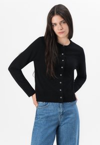 Černý pletený cardigan s kulatým výstřihem, dlouhými rukávy a zapínáním na knoflíky. V kombinaci se světlými modrými džíny.