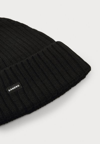 Juodas megztas beanie tipo skrybėlė su šonkauliuota tekstūra ir sulenkta apatine dalimi. Turi mažą baltą etiketę su prekės ženklu "SANDRO."