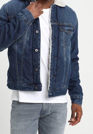 Mężczyzna w niebieskiej dżinsowej kurtce z białą podszewką sherpa, na białej koszulce i szarych dżinsach, poprawiający kołnierz kurtki lewą ręką.