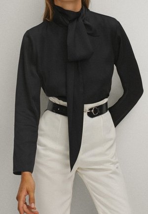 Personne portant un chemisier à manches longues noir avec un nœud à l'avant et un pantalon blanc taille haute, accessoirisé d'une ceinture noire avec une boucle argentée.