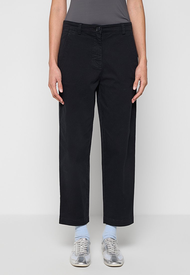 Schwarze Baumwolltwill-Hosen mit geradem Bein, Fronttaschen und einem Knopfverschluss an der Taille; kombiniert mit silbernen Sneakers.