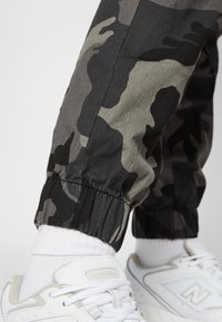 Pantalones jogger de camuflaje en negro, gris y caqui con puños elásticos, combinados con zapatillas New Balance blancas y calcetines blancos texturizados.