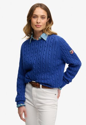 Kvinde iført en blå kabelstrikket sweater over en blåstribet skjorte, hvide bukser og et brunt flettet bælte, poserer med den ene hånd i lommen.