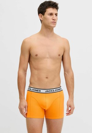 Junger Mann steht und trägt leuchtend orangefarbene Boxershorts mit "JACK & JONES"-Bund vor einem einfarbigen hellen Hintergrund.