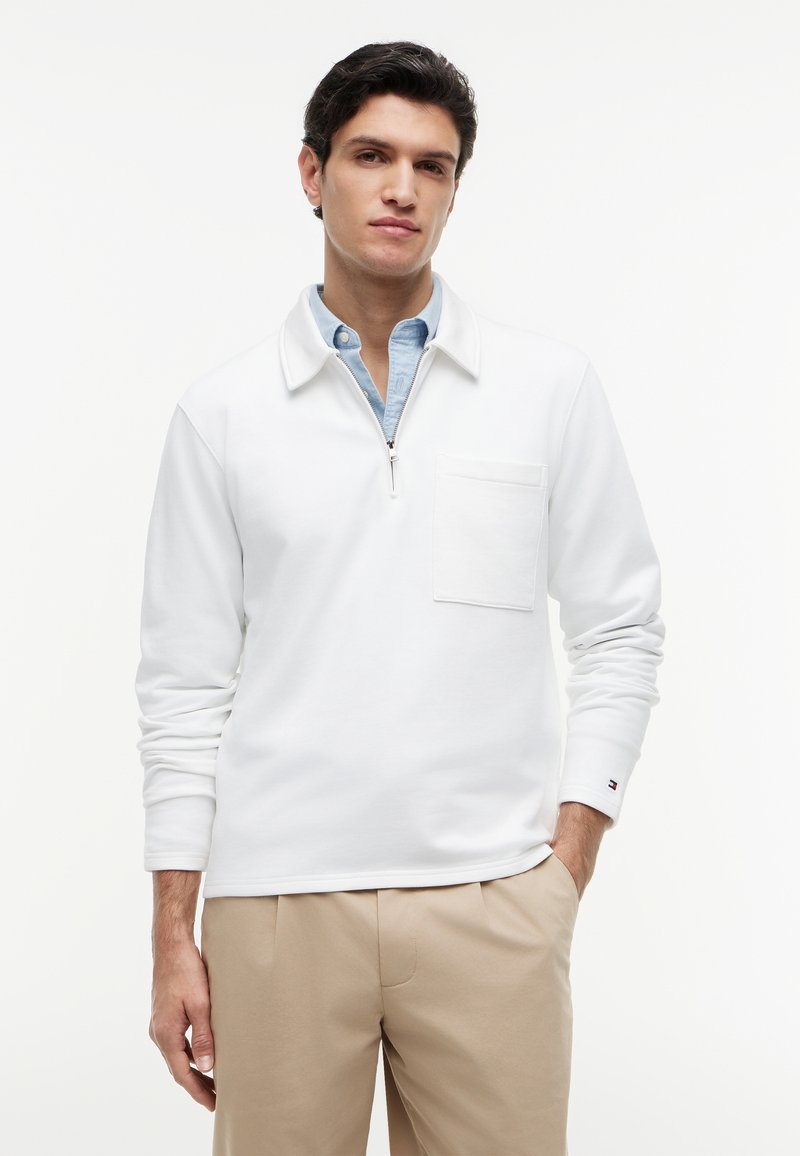 Homme portant un pantalon beige, une chemise bleu clair à col et un pull blanc à manches longues avec une poche poitrine et une fermeture éclair au quart.