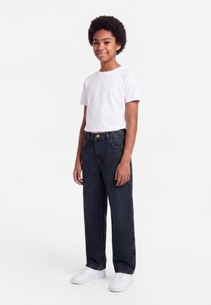Jeune personne debout et souriante portant un t-shirt blanc, un jean foncé et des baskets blanches sur un fond uni clair.