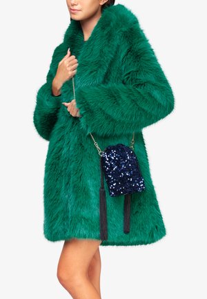 Neppenvacht groene jas met een brede kraag en lange mouwen; gecombineerd met een kleine marineblauwe crossbody tas van pailletten met franjes.