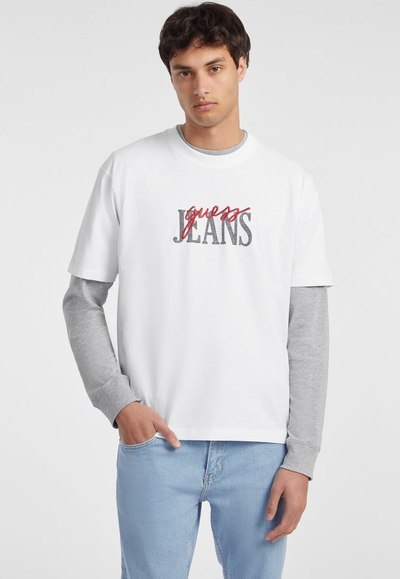 Camiseta blanca de manga corta con gráfico "guess JEANS" en rojo y gris. Lleva una camiseta de manga larga gris debajo. Corte casual.