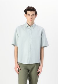 Chemise à manches courtes boutonnée en tissu vert menthe clair, avec un col classique, des boutons à l'avant et une coupe décontractée. Associée à un pantalon vert olive.