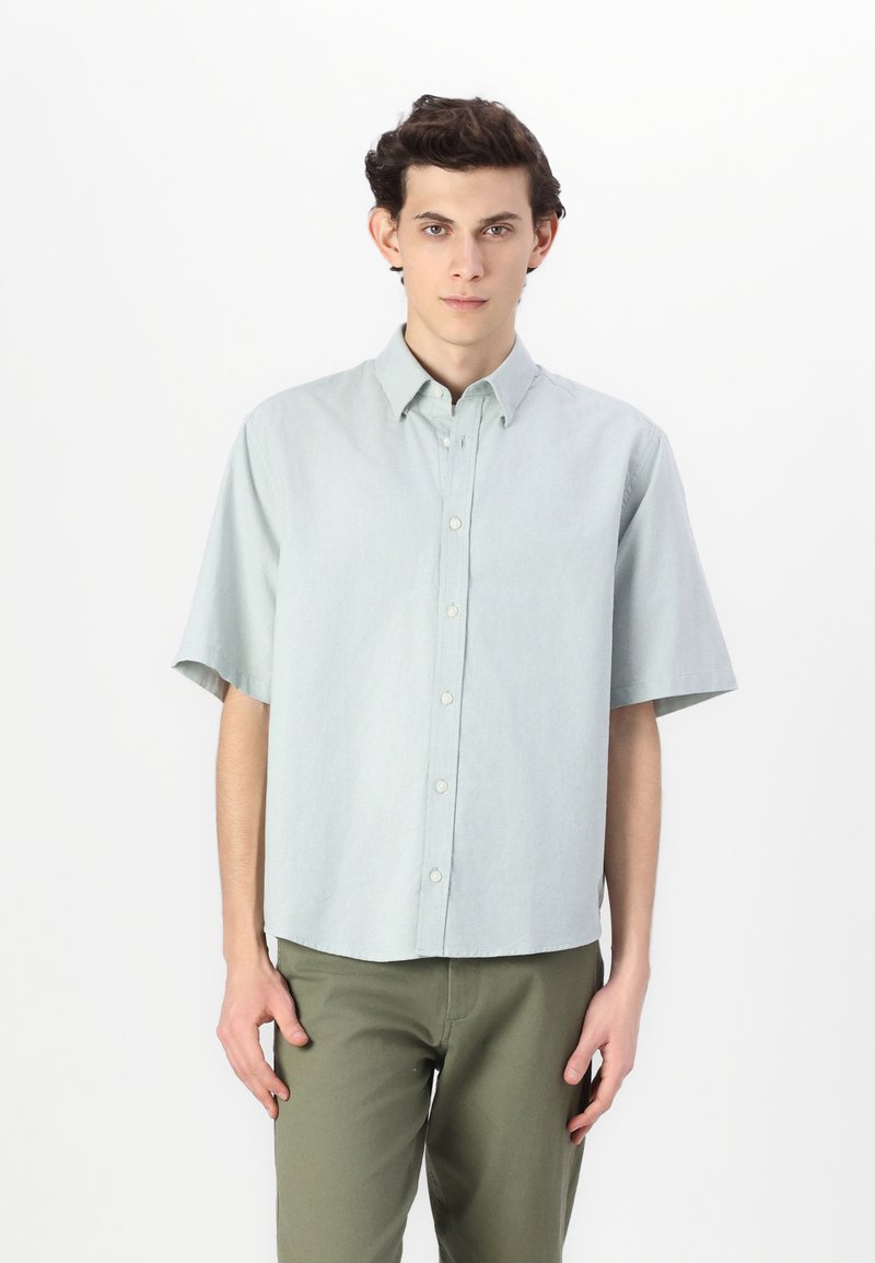 Chemise à manches courtes boutonnée en tissu vert menthe clair, avec un col classique, des boutons à l'avant et une coupe décontractée. Associée à un pantalon vert olive.