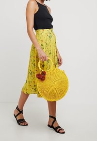 Żółta okrągła pleciona torba o teksturowanym wykończeniu, z okrągłym uchwyt i kolorowymi pomponami, w połączeniu z kwiecistą żółtą spódnicą.