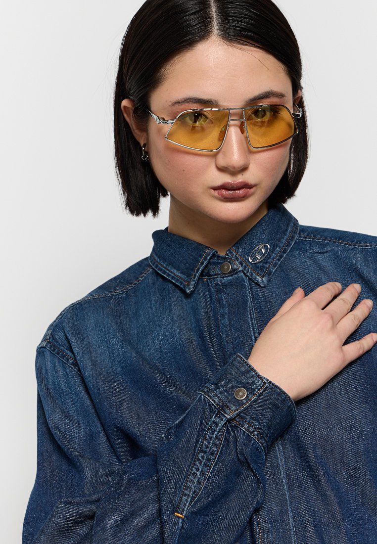 Chemise en denim avec col, fermeture à boutons et manches longues ; portée avec des lunettes de soleil teintées jaunes ; texture lisse, couleur bleu profond et coupe décontractée.