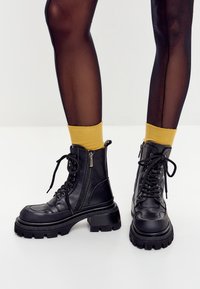 Gambe che indossano collant neri trasparenti, calzini gialli senape e robusti stivali di pelle nera con lacci e zip laterali su un pavimento bianco.