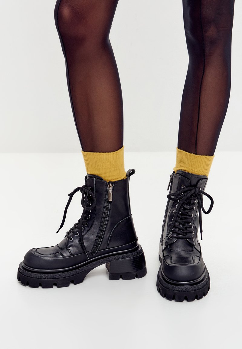 Gambe che indossano collant neri trasparenti, calzini gialli senape e robusti stivali di pelle nera con lacci e zip laterali su un pavimento bianco.