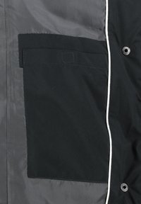 Schwarze Jacke mit einer großen Tasche auf der linken Seite, kontrastierendem grauem Stoff und weißen Paspeln entlang der Kanten sowie Druckknopfdetails.