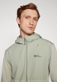 Giacca a vento verde chiaro con cappuccio realizzata in tessuto morbido. Presenta una chiusura con zip sul davanti e un logo sul petto, con un design sobrio e minimale.