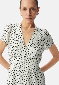 Abito a pois in bianco sporco e nero, con scollatura a V profonda, maniche corte a palloncino e chiusura con bottoni sul davanti, dotato di pince al busto.