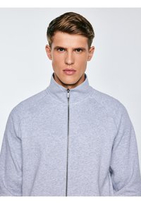 Młody mężczyzna z krótkimi jasnobrązowymi włosami, ubrany w jasnoszary rozpinany sweter z zamkiem, stojący przodem na jednolitym jasnym tle.