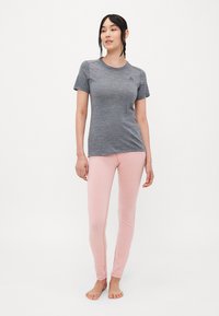 Kvinne med flettehår ikledd en tettsittende grå t-skjorte med korte ermer og lyserosa leggings, som står barfot mot en hvit bakgrunn.