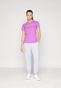 Vijolična bombažna majica s belim napisom na sprednji strani, v kombinaciji s svetlo sivo trenirka in belimi športnimi čevlji, stoji proti enobarvni ozadju.