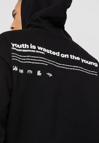 Personne portant un sweat à capuche noir avec un texte blanc au dos indiquant « la jeunesse est gâchée sur les jeunes » et une citation de George Bernard Shaw.