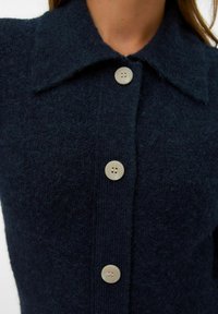 Marineblauwe wollen cardigan met een puntkraag, vijf lichtgekleurde knopen en een getextureerde afwerking; heeft een geribde zoom en manchetten.
