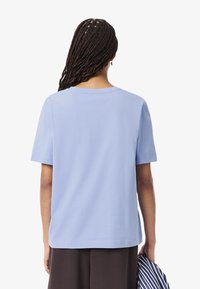 Persona con lunghi capelli intrecciati che indossa una camicia a maniche corte azzurro chiaro e tiene un indumento piegato a strisce al suo fianco.