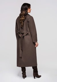 Brun lang jakke lavet af tekstureret stof, med bælte i taljen og vide ærmer. Kombineret med mørke knælange støvler.