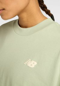 Acercamiento de una persona que lleva una camisa verde claro con el logo de New Balance en el pecho y el cabello trenzado atado con una banda negra.