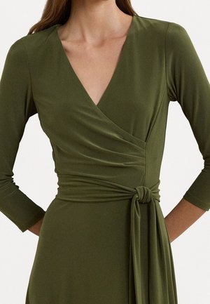 Femme portant une robe portefeuille vert olive avec des manches trois-quarts et une ceinture nouée à la taille, montrée des épaules aux hanches.