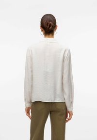 Camicia leggera in crema con una finitura testurizzata, maniche lunghe e dettagli con polsini spaccati, abbinata a pantaloni verde oliva a vita alta.