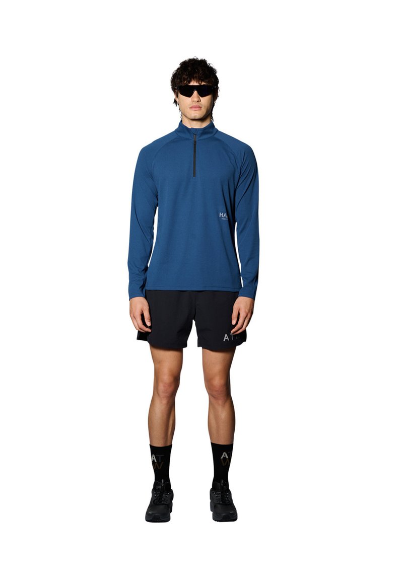 Blå atletisk långärmad tröja med dragkedja vid kragen, svarta shorts, svarta strumpor med logotyp och svarta skor. Modellen står med armarna längs sidorna.