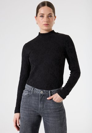 Haut à manches longues noir avec un motif floral texturé, un col montant et une silhouette ajustée, associé à un jean gris taille haute.