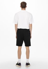Shorts in pile neri con una tasca posteriore, abbinati a una maglietta bianca a maniche corte. Il modello è in piedi di spalle su uno sfondo neutro.