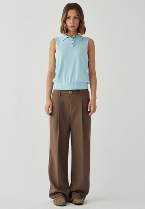 Femme debout de face, portant un haut sans manches bleu clair avec col, un pantalon large marron ceinturé et des mocassins marron.