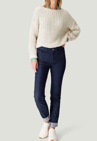 Suéter de punto color crema con un ajuste holgado, combinado con jeans ajustados de color azul oscuro doblados en los puños y zapatillas blancas. Acento verde en los puños.