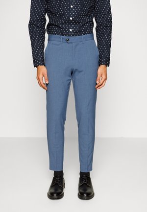 Mann trägt schmal geschnittene hellblaue Anzughose, schwarze Businessschuhe und ein schwarzes, gemustertes Langarmhemd, steht vor einem schlichten Hintergrund.