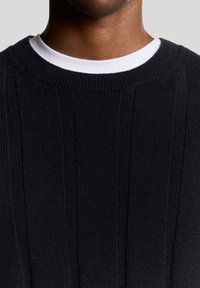 Schwarzer, gestrickter Pullover mit geripptem Muster und rundem Ausschnitt, über einem weißen Hemd getragen. Nahaufnahme zeigt Details des Stoffes.