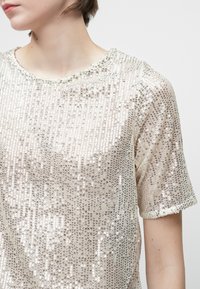 Top de manga corta en color beige claro con una superficie texturizada y con lentejuelas. Presenta un escote redondo y una calidad ligeramente transparente.