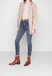 Beige konstläder blazer med en strukturerad yta, kombinerad med en figursydd krämfärgad crop top, mörkblå skinny jeans och bruna lackade ankelstövlar.