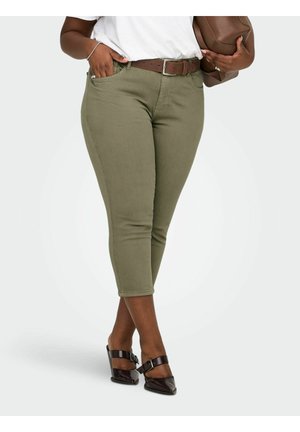 Osoba nosiaca krátke olivovo zelené nohavice s hnedým opaskom, drží hnedú tašku a má na sebe tmavohnedé topánky na podpätku.