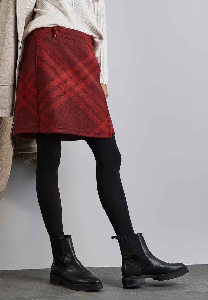 Femme portant une jupe en laine à carreaux couleur bordeaux, des collants noirs, des bottines en cuir noires et un pull côtelé crème sur fond gris.