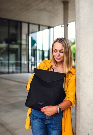 Schwarzer Rucksack mit Klappendesign, aus Stoff gefertigt, mit einem auffälligen Logo an der Unterseite. Getragen von einer Person in einer gelben Jacke.