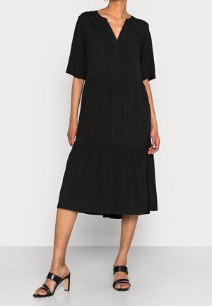 Femme portant une robe midi noire ample à manches courtes et col en V, associée à des sandales à talons noires à deux brides.